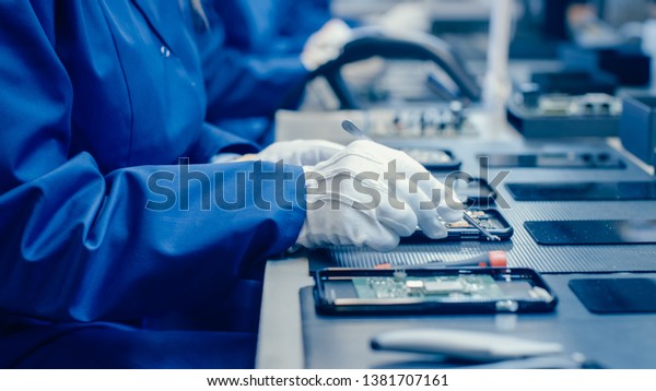 Close Up of a Female Electronics Factory\
Worker in Blue Work Coat and Protective Glasses Assembling\
Smartphones with Screwdriver. High Tech Factory Facility with more\
Employees in the\
Background.