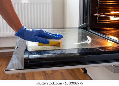 Close Up Of Female Hand With Blue Protective Gloes Cleaning Oven.Woman Hand Cleaning Dirty Oven Door In The Kitcken.