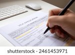 Close up of a female hand filling out a visa application document, white table with computer keyboard and a mouse in background