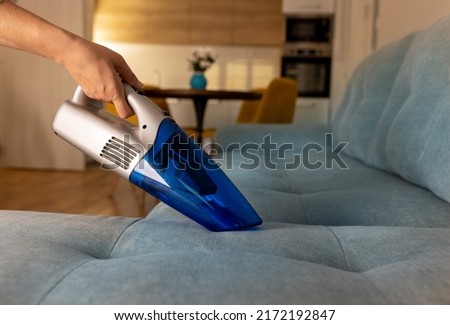 Close up of female hand with gloves holding handy rechargeable wireless vacuum cleaner hooving hair and crumbs from sofa in living room in background . Housekeeping, cleaning concept