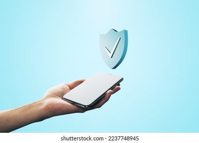 Close up of female hand holding mobile phone with creative antivirus shield icon on light blue background. Secure phone usage, protection and web safety concept