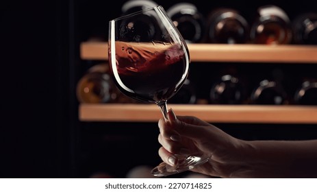 Close up female hand swirling red wine in wine glass. Wine expert tasting, rating and drinking wine, bottles in background. - Shutterstock ID 2270714825
