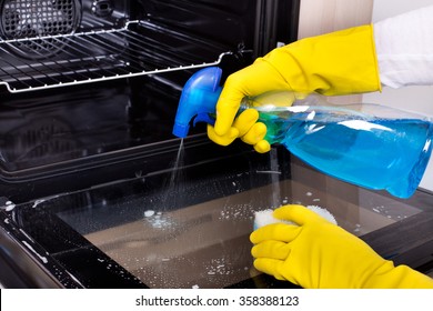 Close Up Of Female Hand With Yellow Protective Gloves Spraying Detergent From Bottle On The Oven Door