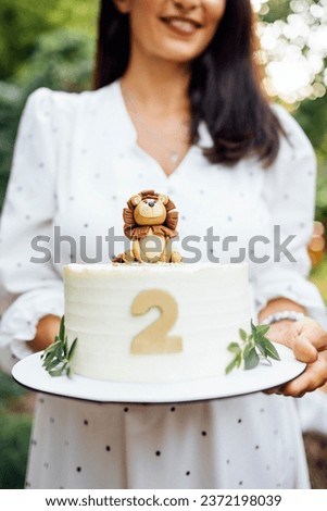 Close up of female hands hold a birthday baby cake. Delicious sweet pie with cute lion cub, number two and leaves. Smiling brownhaired woman in white dress on the background. Cooking and kids party. Stock photo © 