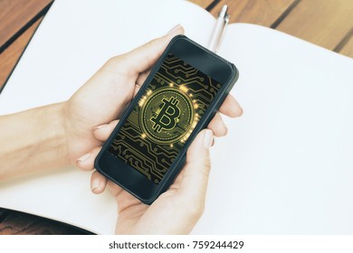 Close up of female hands holding mobile phone with digital bitcoin on screen at wooden desk with empty copybook. Online banking concept