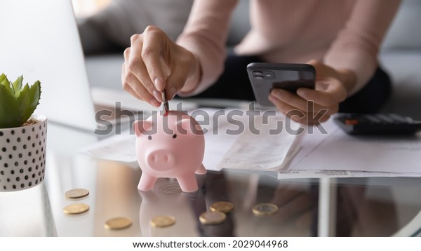 Close up female hands holding smartphone put euro\
coin into piggy bank, advanced modern tech user using of mobile\
budget tracking app for personal finances, family money management,\
e-banking concept