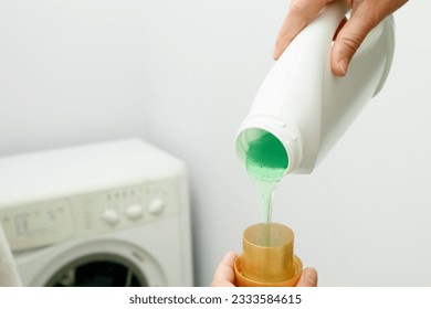 Close up of female hands pouring liquid laundry detergent into cap in the washing machine. Pour green washing liquid, wash machine blured in background. - Shutterstock ID 2333584615