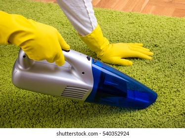 Close up of female hands with rubber gloves holding cordless vacuum cleaner and hoovering green carpet on parquet floor