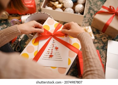 Close up female hands tying a red ribbon bow on a craft gift box. Woman wrapping a Christmas gift while sitting on the carped in the living room. - Powered by Shutterstock