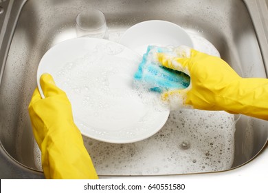 Close up of female hands in yellow protective rubber gloves washing white plate with blue cleaning sponge against kitchen sink full of foam and tableware 