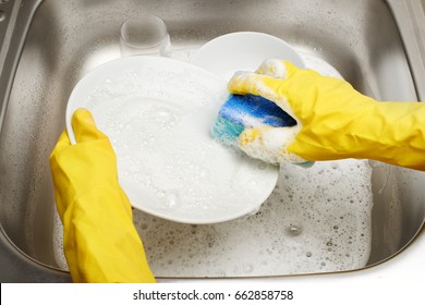 Close up of female hands in yellow protective rubber gloves washing white plate with blue cleaning sponge against kitchen sink full of foam and tableware  - Powered by Shutterstock