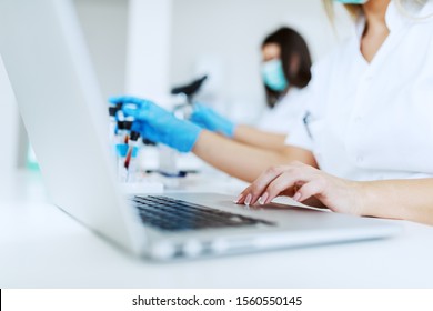 Close up of female lab assistant in white uniform sitting in lab and using laptop for data entry. In background is her colleague working.