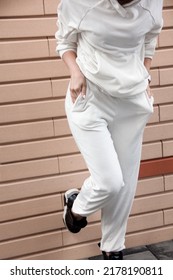 Close Up Female Leg, Teen Walking Look. Young Girl In White Oversize Suit Is Standing Casual With Hands In Pockets On The Beige Brick Street Building Background. Lifestyle Concept, Free Space