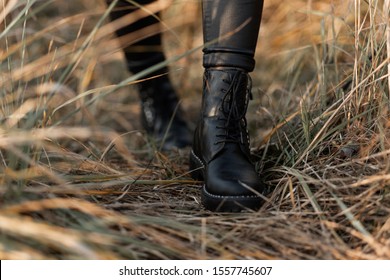 Close up of female legs in fashionable leather black boots. Trendy girl in stylish leggings in fashion shoes walks on the dry grass on the street. Casual autumn style.
