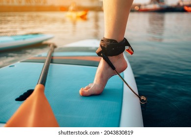 Close Up Of Female Legs Standing On Blue Yellow Color SUP Stand Up Paddle Board Or Surfboard Puts On Ankle Leash At Sunset. Active Family Summer Vacation Time Sport