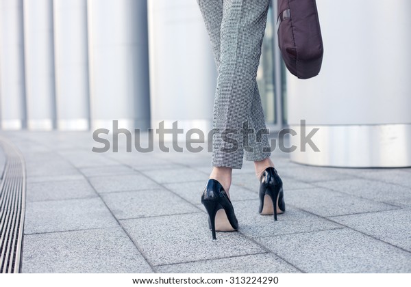 Close up of female legs\
of worker standing near her office. The woman is wearing formalwear\
and shoes on high heels. She is holding a handbag. Copy space in\
left side