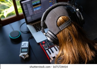 Close Up Of A Female Music Producer With Headphones In Her Home Studio, Desk With Digital Recorder, Smart Home Assistant, Notebook And A Midi Keyboard. Window With Nature In The Background.