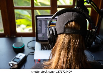 Close Up Of A Female Music Producer With Headphones In Her Home Studio, Desk With Digital Recorder, Smart Home Assistant, Notebook And A Midi Keyboard. Window With Nature In The Background.