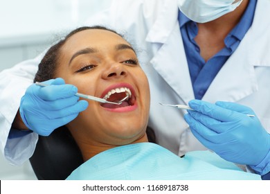 Close Up Of Female Patient Being Examined By Dentist, Wearing In Latex Gloves, In Clinic. Young Afro Woman, Sitting In Chair Of Dentist, Looking Straight With Open Mouth While Doctor Checking Teeth.