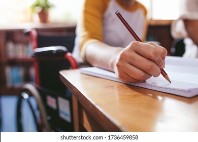 Close up of female student taking notes sitting in wheelchair at desk. Focus on hand writing in notebook with a pencil. - Powered by Shutterstock