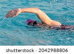 Close up of a female swiming in a pool with her arm raised in the air swimming freestyle wearing a pink cap and googles.