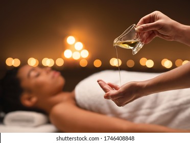 Close up of female therapist applying massage oil on hands before therapy over black lady lying on massage table