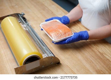 Close Up Of Female Worker In Factory Or Shop Holding Piece Of Salmon Wrapped In Food Film. Woman In Blue Gloves Holds Salmon Fillet Lying On White Tray Near Large Roll Of Film Lying On Table.