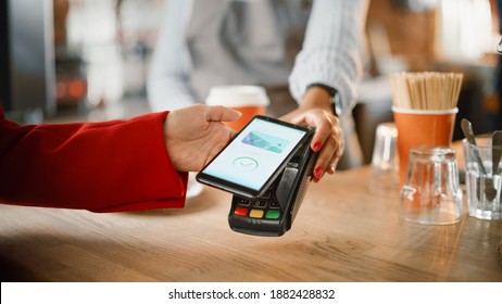 Close Up of a Feminine Hand Holding a Smartphone with an NFC Payment Technology Used for Paying for Take Away Coffee in a Cafe. Customer Uses Mobile to Pay for Latte Through a Credit Card Terminal. - Powered by Shutterstock