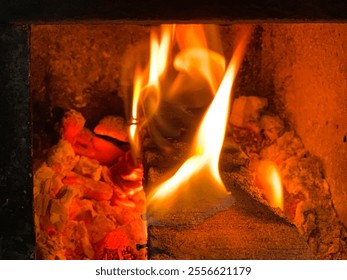 Close up of fire flames from a burning piece of wood inside of traditional stove, the end of the wood has blackened into charcoal. Embers and ashes lie around. - Powered by Shutterstock