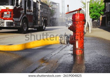 Close up of fire hydrant with hose attached and defocused fire truck. Supply line or hydrant line width large diameter. Water spaying from hole. Fire hall responding to fire in city. Selective focus.
