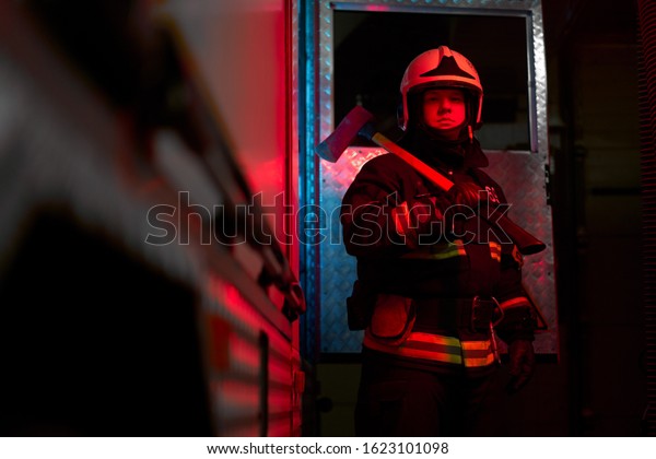 Close up firefighter man with hammer on his\
shoulder standing near fire\
engine