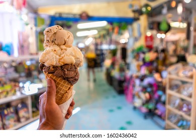 A Close Up And First Person Perspective Of A Man Holding A Waffle Cone With Two Scoops Of Ice Cream At A Shop, With A Colorful Store In The Background