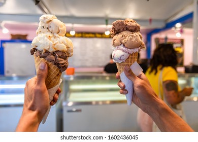 A Close Up And First Person Perspective Of A Man Holding Two Waffles Cone With Scoops Of Ice Cream At A Shop, With Blurry Menu In The Background