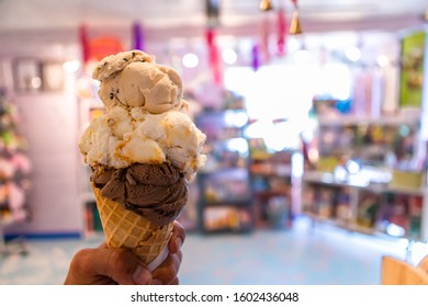 A Close Up And First Person Perspective Of A Man Holding A Waffle Cone With Two Scoops Of Ice Cream At A Shop, With A Colorful Store In The Background