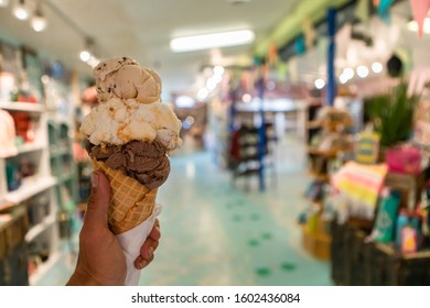 A Close Up And First Person Perspective Of A Man Holding A Waffle Cone With Two Scoops Of Ice Cream At A Shop, With A Colorful Store In The Background