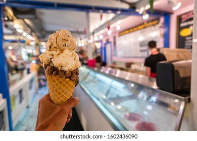 A Close Up And First Person Perspective Of A Man Holding A Waffle Cone With Scoops Of Ice Cream At A Shop, With Blurry Counter In The Background