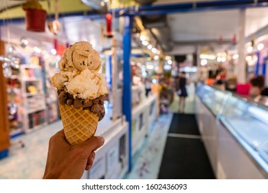 A Close Up And First Person Perspective Of A Man Holding A Waffle Cone With Scoops Of Ice Cream At A Shop, With Blurry Counter In The Background