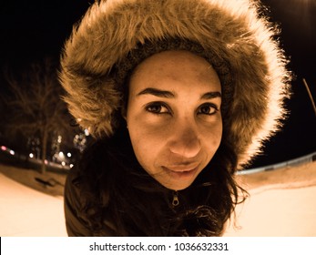 Close Up Fish Eye Wide Angle Photograph Of A Beautiful Young African American Woman With Dark Long Hair Wearing A Coat With A Furry Lined Hood Over Her Head And Looking Into The Camera.