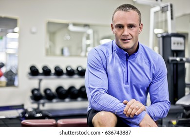 Close Up Of Fitness Trainer With Blue Shirt, Inside Room Of Luxury Gym, Heavy Exercise Tools Near Wall, A Body Builder Sitting On A Bench, Perfect Lights And Designs, He Is So Healthy.