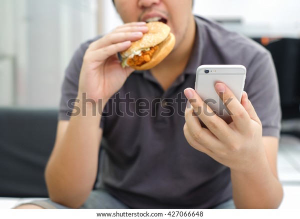 アジア人男性の手でスマートフォンを演奏し 家きん類や野菜のコンセプトの中でハンバーガーを食べるようにします の写真素材 今すぐ編集