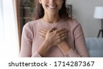 Close up focus on happy sincere female holding folded hands on chest. Emotional positive kind candid millennial woman feeling thankful indoors, showing gratitude sign, believe faith charity concept.