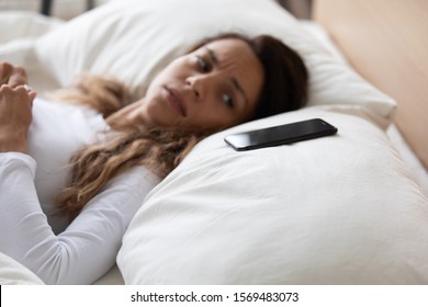 Close up focus on mobile phone is on pillow, frightened woman lying in bed looking on smartphone feels afraid and scared, concept of cyberbullying or stalking, victim feels fear from call or message