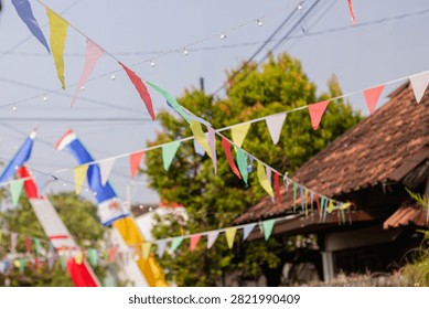 Close focus on multicolored triangular pennant flags hanging outdoors with a traditional village house roof and lush green trees in the background. High Quality Photo