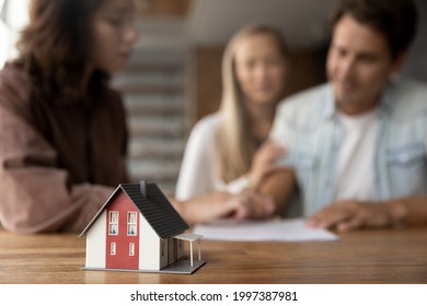 Close up focus on small house model standing on table with blurred couple clients signing contract agreement with real estate agent, purchasing own dwelling apartment, professional service concept.