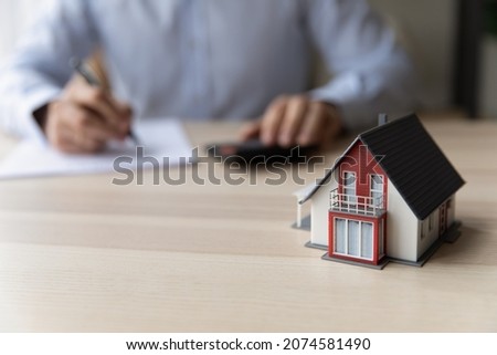 Close up focus on small house model with young man handwriting notes in paper organizer, planning mortgage payments, calculating expenditures for real estate investment or purchasing own accommodation