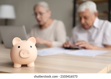 Close Up Focus On Small Piggybank On Table With Blurred Middle Aged Old Family Couple Doing Financial Paperwork, Managing Household Budget Or Planning Investment On Background, Retirement Lifestyle.