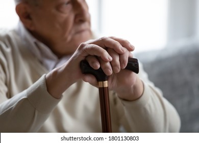 Close up focus on wrinkled male hands leaning at wooden stick. Lost in thoughts upset old male pensioner having problems with walking, suffering from loneliness indoors, retirement lifestyle concept.