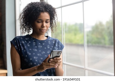 Close up focused African American young woman using smartphone, looking at screen, standing near window at home, chatting or shopping online, searching information, reading news in message - Powered by Shutterstock