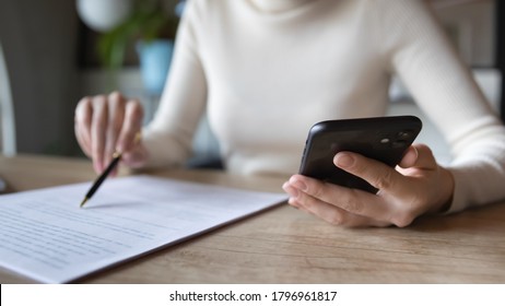Close up focused financier reading paper contract agreement, checking latest investment news in smartphone application indoors, concentrated female editor reading document or report at workplace.