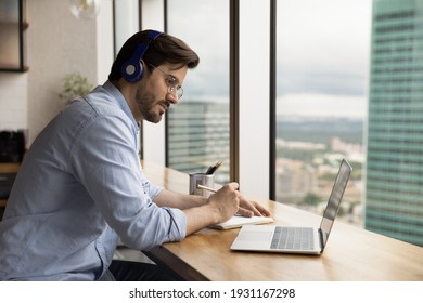 Close up focused man wearing headphones and glasses using laptop, taking notes, intern or student watching webinar, training, listening to lecture, sitting at desk near window, studying online - Powered by Shutterstock